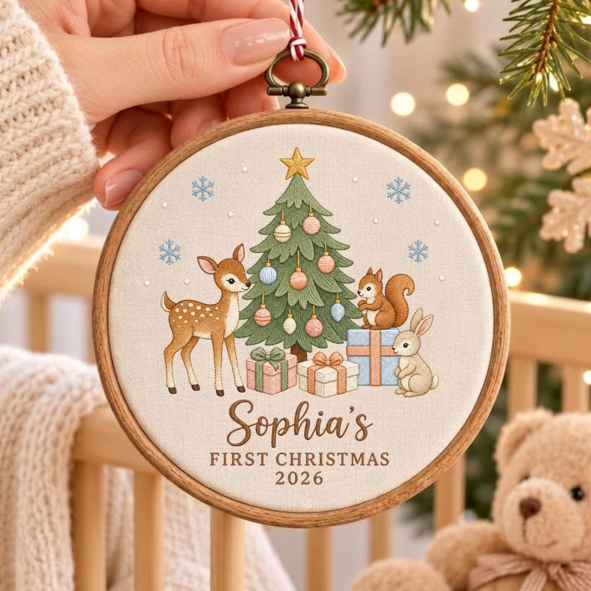 May include: A round wooden embroidery hoop ornament featuring a fabric center with an embroidered Christmas tree, a deer, a squirrel, and a rabbit. The text reads Sophia's FIRST CHRISTMAS 2026. The design includes colorful gift boxes and snowflakes.