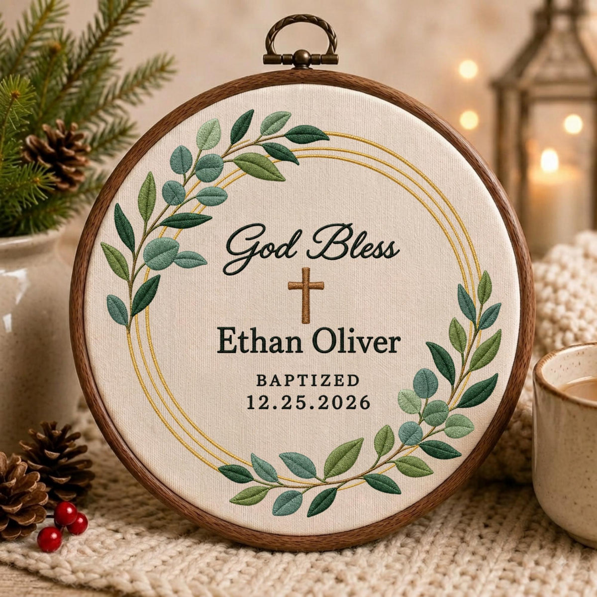 May include: A circular wooden embroidery hoop containing off-white fabric with embroidered green eucalyptus leaves, a brown cross, and black text reading God Bless Ethan Oliver BAPTIZED 12.25.2026, framed by thin gold circular stitched lines.