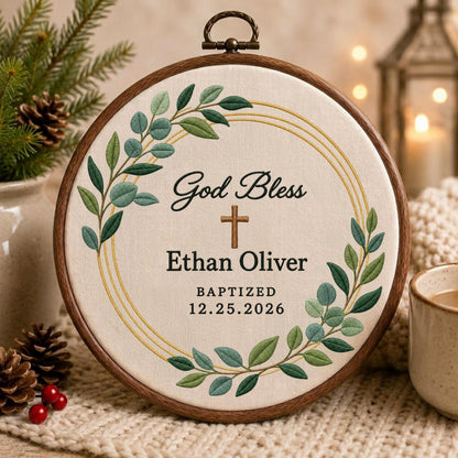 May include: A circular wooden embroidery hoop containing off-white fabric with embroidered green eucalyptus leaves, a brown cross, and black text reading God Bless Ethan Oliver BAPTIZED 12.25.2026, framed by thin gold circular stitched lines.