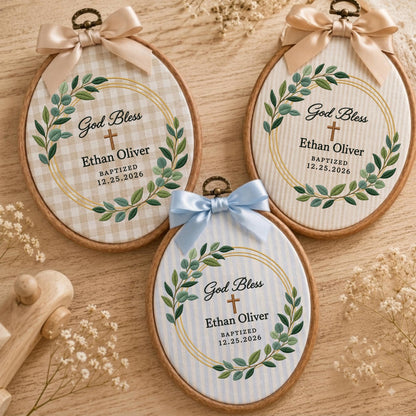 May include: Three oval wooden embroidery hoops featuring fabric backgrounds with gingham or striped patterns. Each hoop is topped with a satin ribbon bow and displays a printed wreath of green leaves, a cross, and the text God Bless Ethan Oliver.