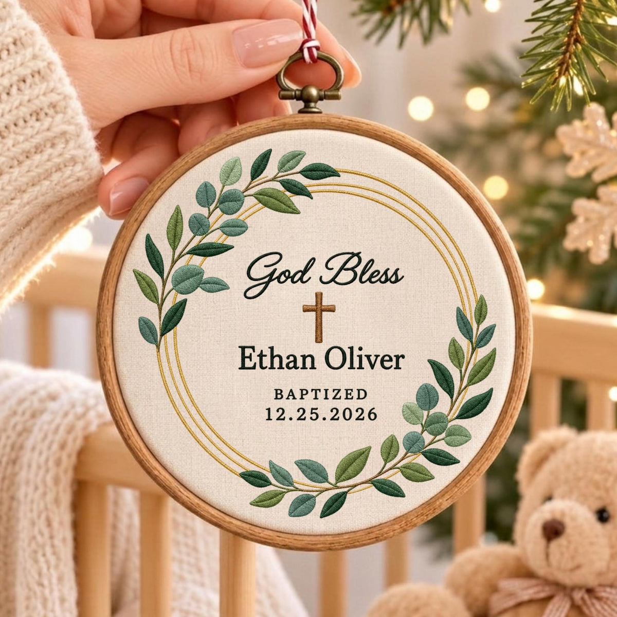 May include: A circular wooden embroidery hoop containing off-white fabric with green leaf embroidery, a gold circular frame, and a brown cross. The text reads God Bless, Ethan Oliver, BAPTIZED, and 12.25.2026 in black, with a red and white string.