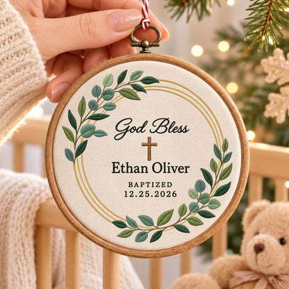 May include: A circular wooden embroidery hoop containing off-white fabric with green leaf embroidery, a gold circular frame, and a brown cross. The text reads God Bless, Ethan Oliver, BAPTIZED, and 12.25.2026 in black, with a red and white string.