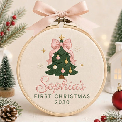 May include: A round wooden embroidery hoop containing cream fabric with an embroidered green Christmas tree, pink bow, and ornaments. The text reads "Sophia's FIRST CHRISTMAS 2030" in pink and green. A large pink satin ribbon is tied to the top.