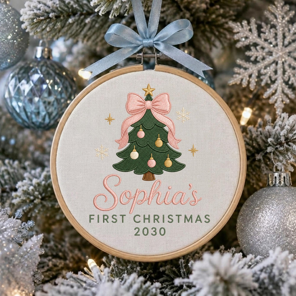 May include: A round wooden embroidery hoop ornament featuring a cream-colored fabric background. The design includes an embroidered green Christmas tree with pink and gold ornaments, topped with a pink bow, and the text Sophia's FIRST CHRISTMAS 2030.