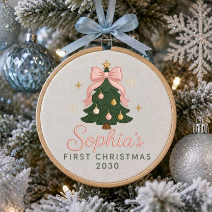 May include: A round wooden embroidery hoop ornament featuring a cream-colored fabric background. The design includes an embroidered green Christmas tree with pink and gold ornaments, topped with a pink bow, and the text Sophia's FIRST CHRISTMAS 2030.