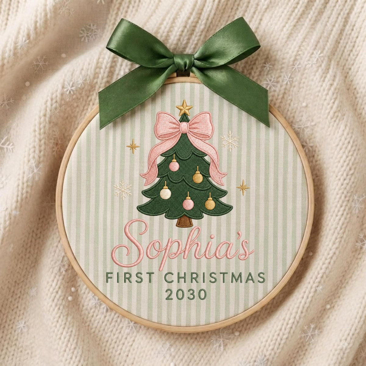 May include: A circular wooden embroidery hoop featuring a light green and white striped fabric background. The design includes an embroidered green Christmas tree with pink and gold ornaments, a pink bow, and the text Sophia's FIRST CHRISTMAS 2030.