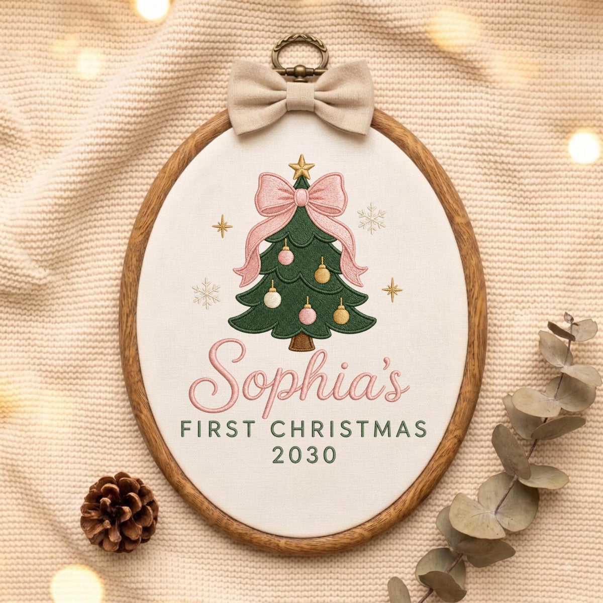 May include: An oval wooden embroidery hoop containing white fabric with an embroidered green Christmas tree, pink bow, and ornaments. The text reads Sophia's FIRST CHRISTMAS 2030. A beige fabric bow is attached to the top of the wooden frame.