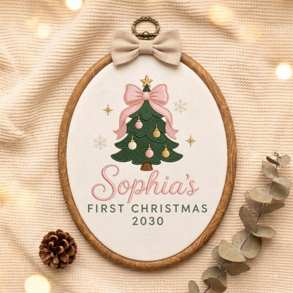 May include: An oval wooden embroidery hoop containing white fabric with an embroidered green Christmas tree, pink bow, and ornaments. The text reads Sophia's FIRST CHRISTMAS 2030. A beige fabric bow is attached to the top of the wooden frame.