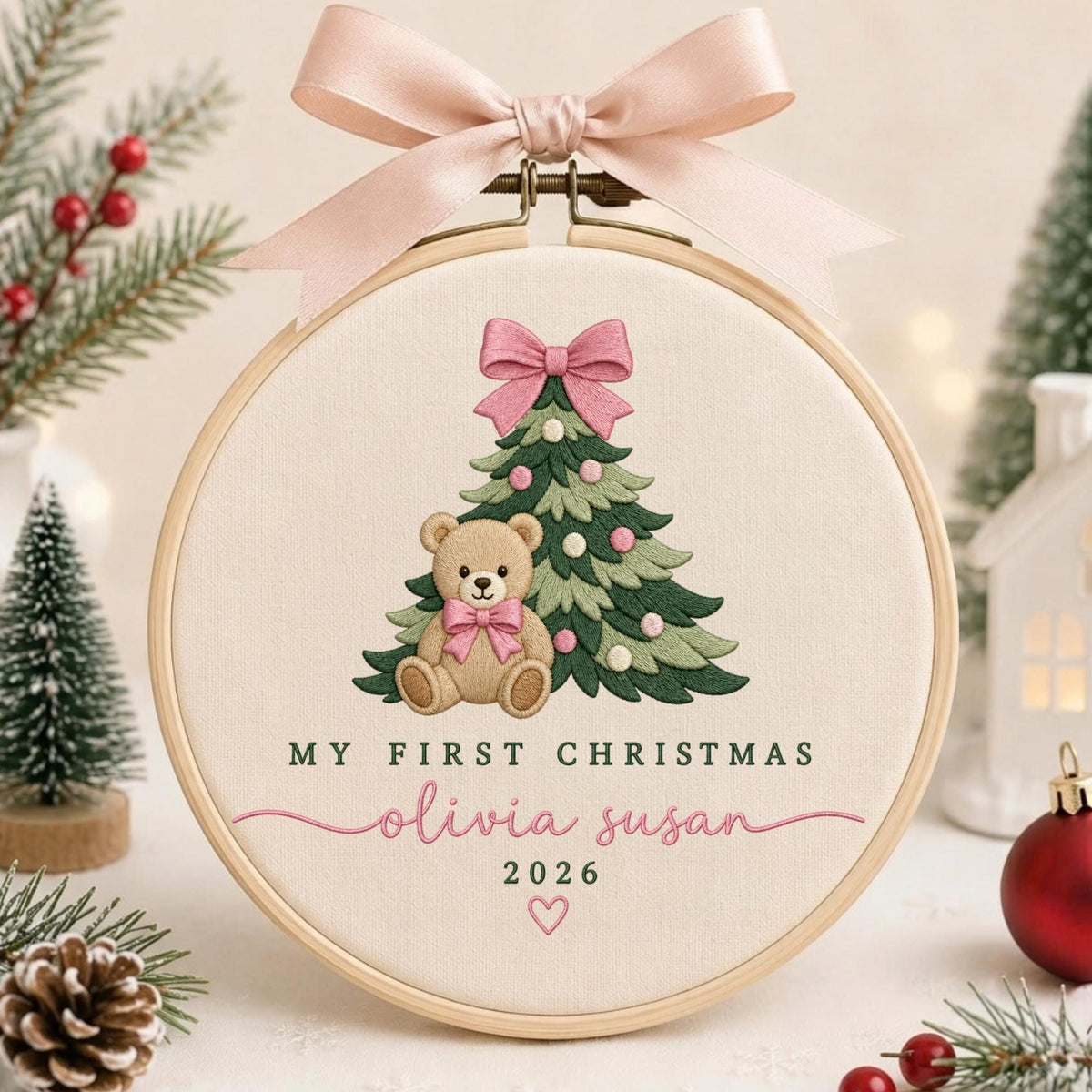May include: A round wooden embroidery hoop containing cream-colored fabric with an embroidered Christmas tree, a teddy bear with a pink bow, and the text "MY FIRST CHRISTMAS", "olivia susan", "2026", and a small heart, topped with a pink satin ribbon.
