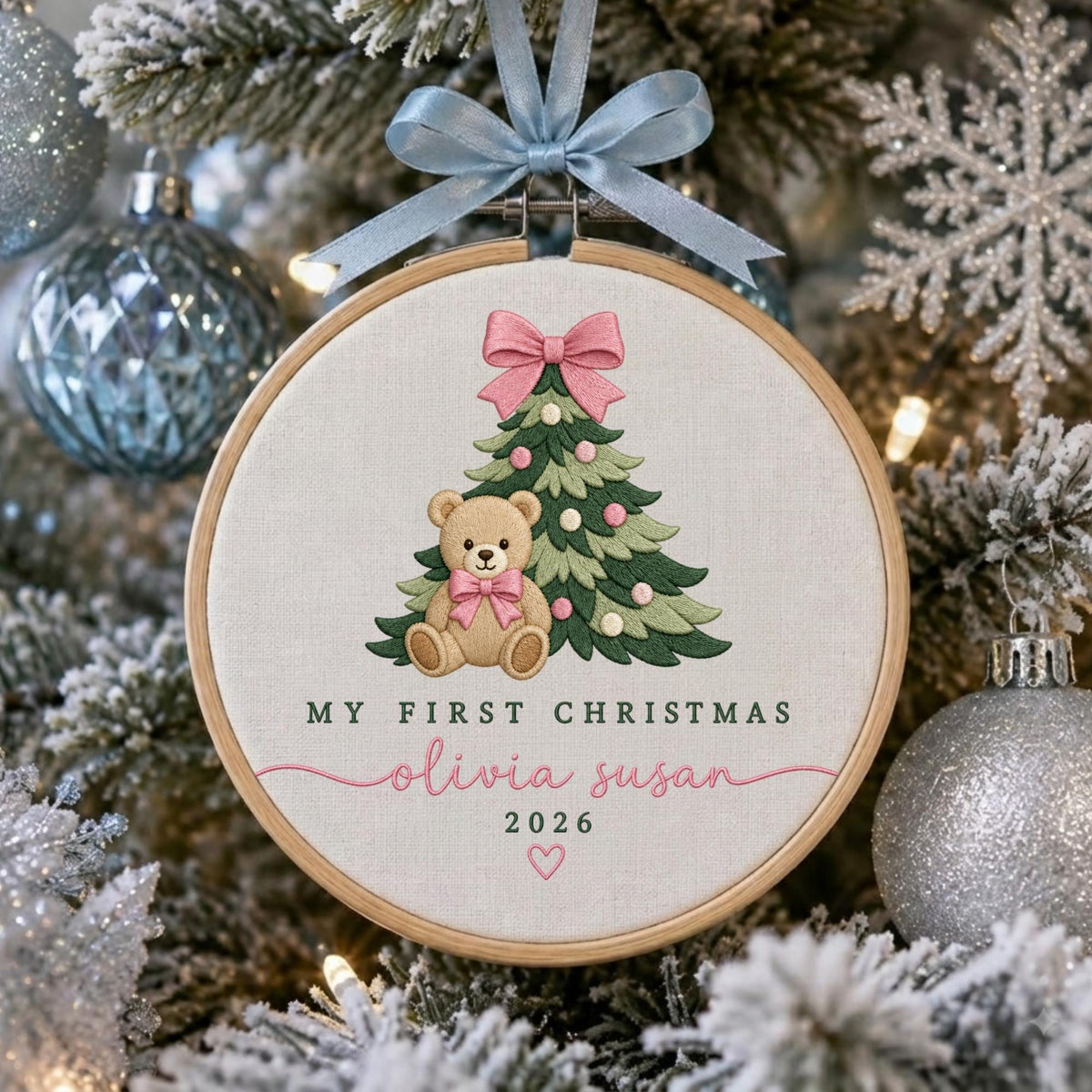 May include: A round wooden embroidery hoop ornament featuring a light-colored fabric background. The design includes an embroidered green Christmas tree with pink ornaments, a brown teddy bear with a pink bow, and the text "MY FIRST CHRISTMAS", "olivia susan", "2026", and a small pink heart. A light blue ribbon is tied at the top.