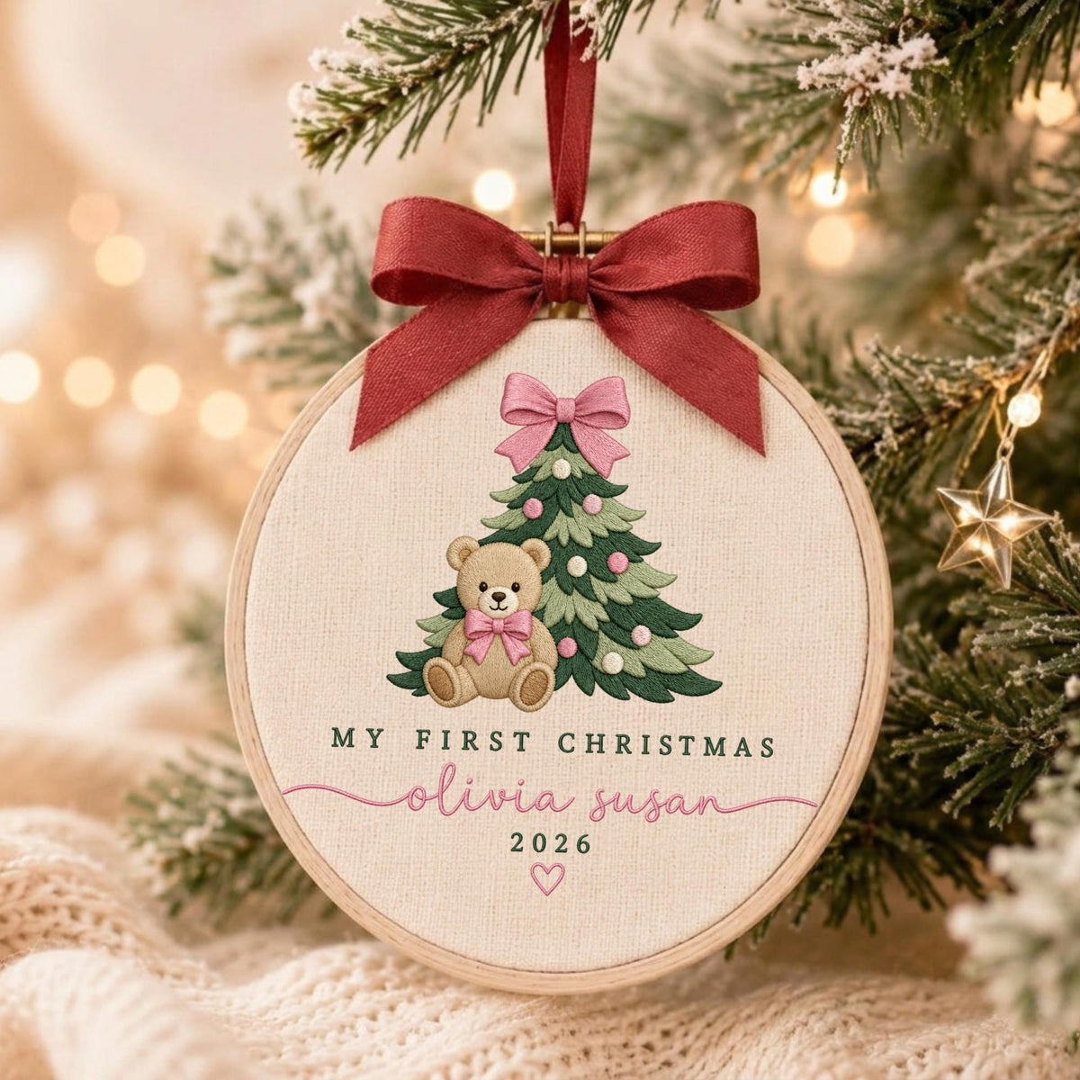 May include: A round embroidery hoop ornament featuring a beige fabric background with a stitched green Christmas tree, a brown teddy bear, and a pink bow. The text reads MY FIRST CHRISTMAS, olivia susan, 2026, with a small heart and a dark red ribbon.