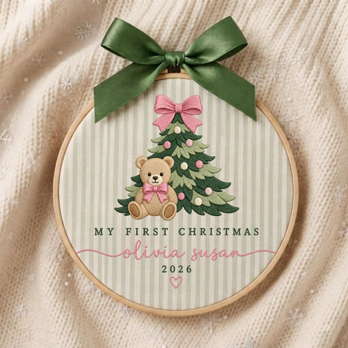 May include: A circular embroidery hoop featuring a light green and white striped fabric. The design includes an embroidered Christmas tree, a teddy bear with a pink bow, and the text "MY FIRST CHRISTMAS olivia susan 2026". A dark green satin ribbon bow is tied at the top.