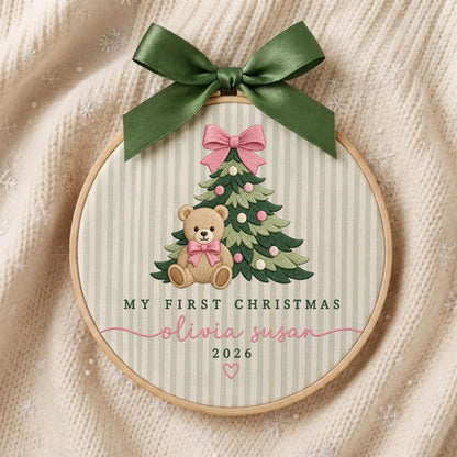 May include: A circular embroidery hoop featuring a light green and white striped fabric. The design includes an embroidered Christmas tree, a teddy bear with a pink bow, and the text "MY FIRST CHRISTMAS olivia susan 2026". A dark green satin ribbon bow is tied at the top.