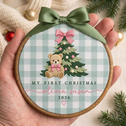 May include: A round wooden embroidery hoop ornament featuring a light green and white gingham fabric background. The design includes an embroidered Christmas tree with a pink bow, a teddy bear, and the text My First Christmas Olivia Susan 2026.