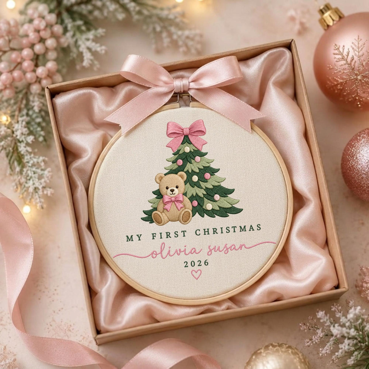 May include: A round wooden embroidery hoop containing a fabric panel with a stitched design of a green Christmas tree, a brown teddy bear, and a pink bow. The text reads MY FIRST CHRISTMAS, olivia susan, 2026, with a small heart icon below.