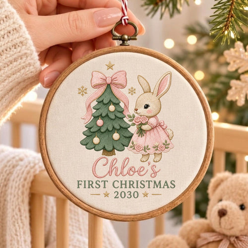 May include: A round wooden embroidery hoop ornament featuring a cream fabric background with a green Christmas tree, a pink bow, and a tan bunny holding flowers. The text reads Chloe's FIRST CHRISTMAS 2030 with small gold stars.
