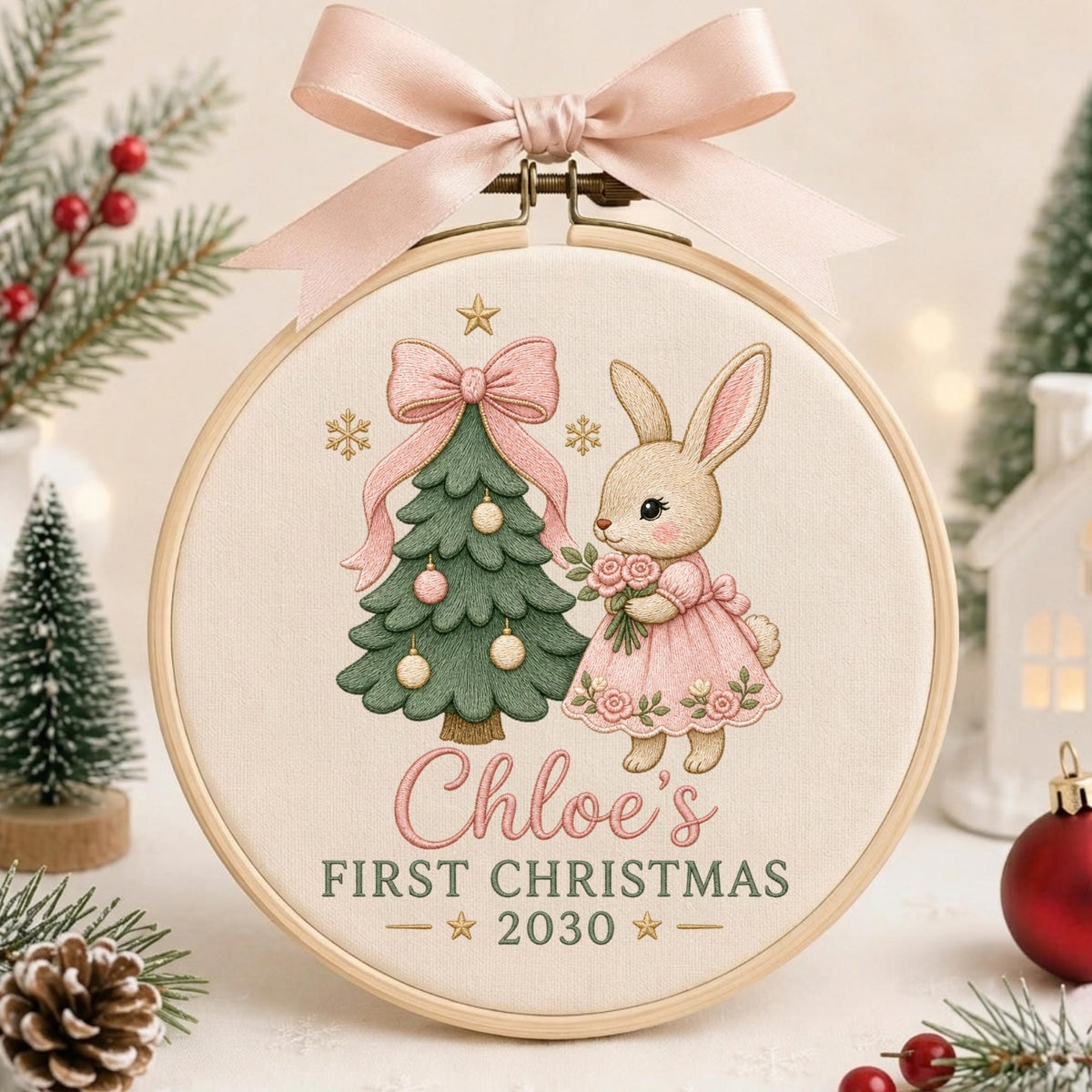May include: A round wooden embroidery hoop featuring a cream-colored fabric with a printed design of a green Christmas tree and a rabbit in a pink dress. The text reads Chloe's FIRST CHRISTMAS 2030. A light pink satin ribbon bow is tied at the top.