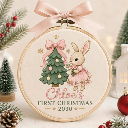 May include: A round wooden embroidery hoop featuring a cream-colored fabric with a printed design of a green Christmas tree and a rabbit in a pink dress. The text reads Chloe's FIRST CHRISTMAS 2030. A light pink satin ribbon bow is tied at the top.