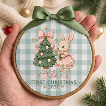 May include: A round wooden embroidery hoop ornament featuring a light green and white buffalo check background. The design includes a decorated Christmas tree, a rabbit in a pink dress, and the text Chloe's FIRST CHRISTMAS 2030. A sage green satin ribbon bow is attached to the top.