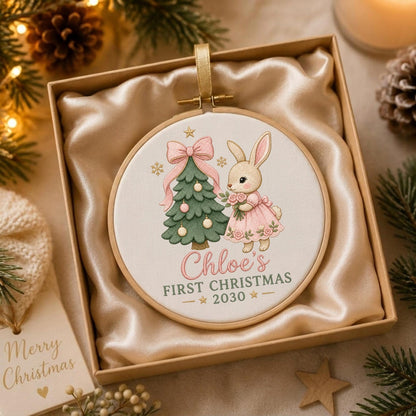 May include: A round wooden embroidery hoop ornament featuring a cream fabric center with a printed illustration of a rabbit in a pink dress next to a decorated green Christmas tree. The text reads Chloe's FIRST CHRISTMAS 2030.