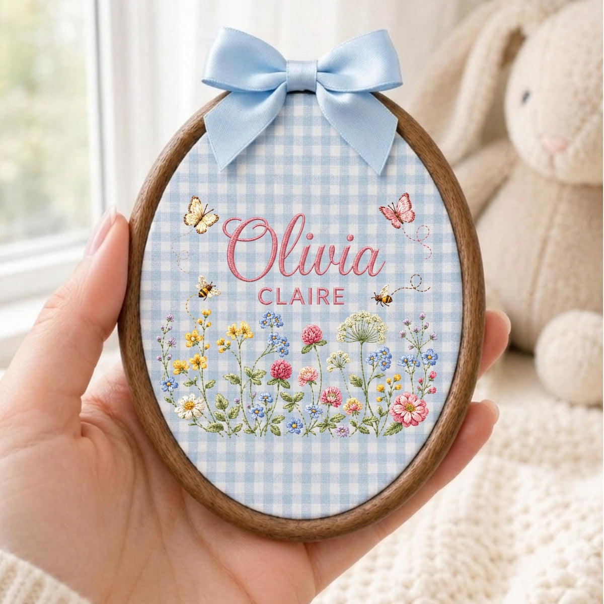 May include: An oval embroidery hoop with a dark wood frame, featuring a light blue and white gingham fabric background. The fabric is embroidered with the names Olivia Claire in pink, surrounded by colorful wildflowers, bees, and butterflies, topped with a light blue satin ribbon bow.