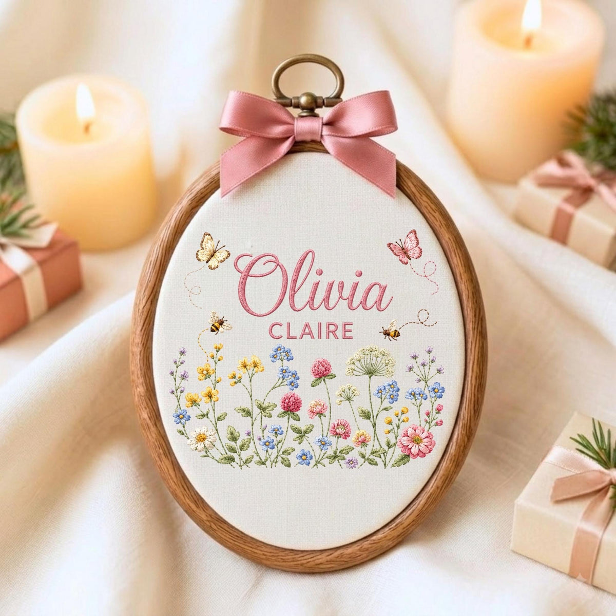 May include: An oval wooden embroidery hoop containing white fabric with the embroidered names Olivia Claire in pink script and block letters. The design features colorful wildflowers, two small bees, and two butterflies, topped with a pink satin bow.