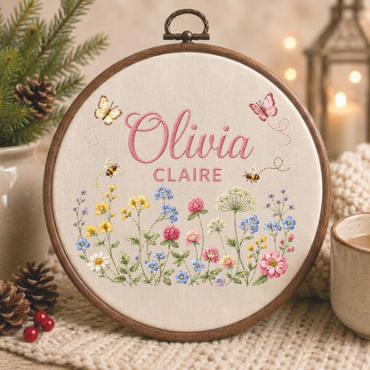 May include: A circular wooden embroidery hoop containing off-white fabric with the embroidered names Olivia Claire in pink script and block lettering. The design features colorful wildflowers, two small bees, and two butterflies in flight.