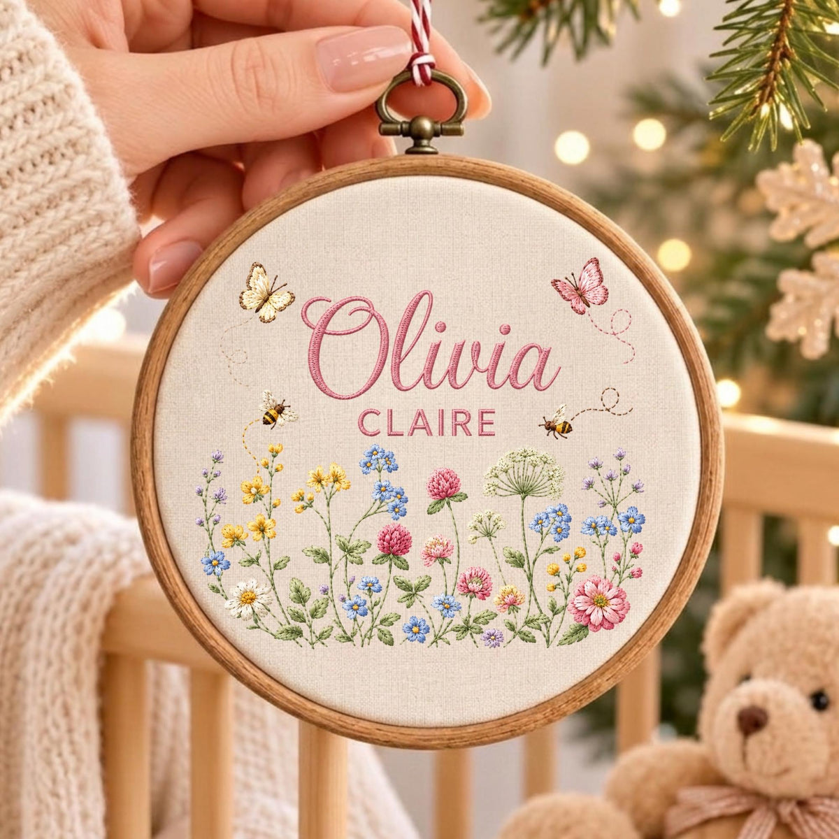 May include: A circular wooden embroidery hoop containing off-white fabric with the embroidered names Olivia Claire in pink script and block letters. The design features colorful wildflowers, two small bees, and two butterflies in pink and yellow.