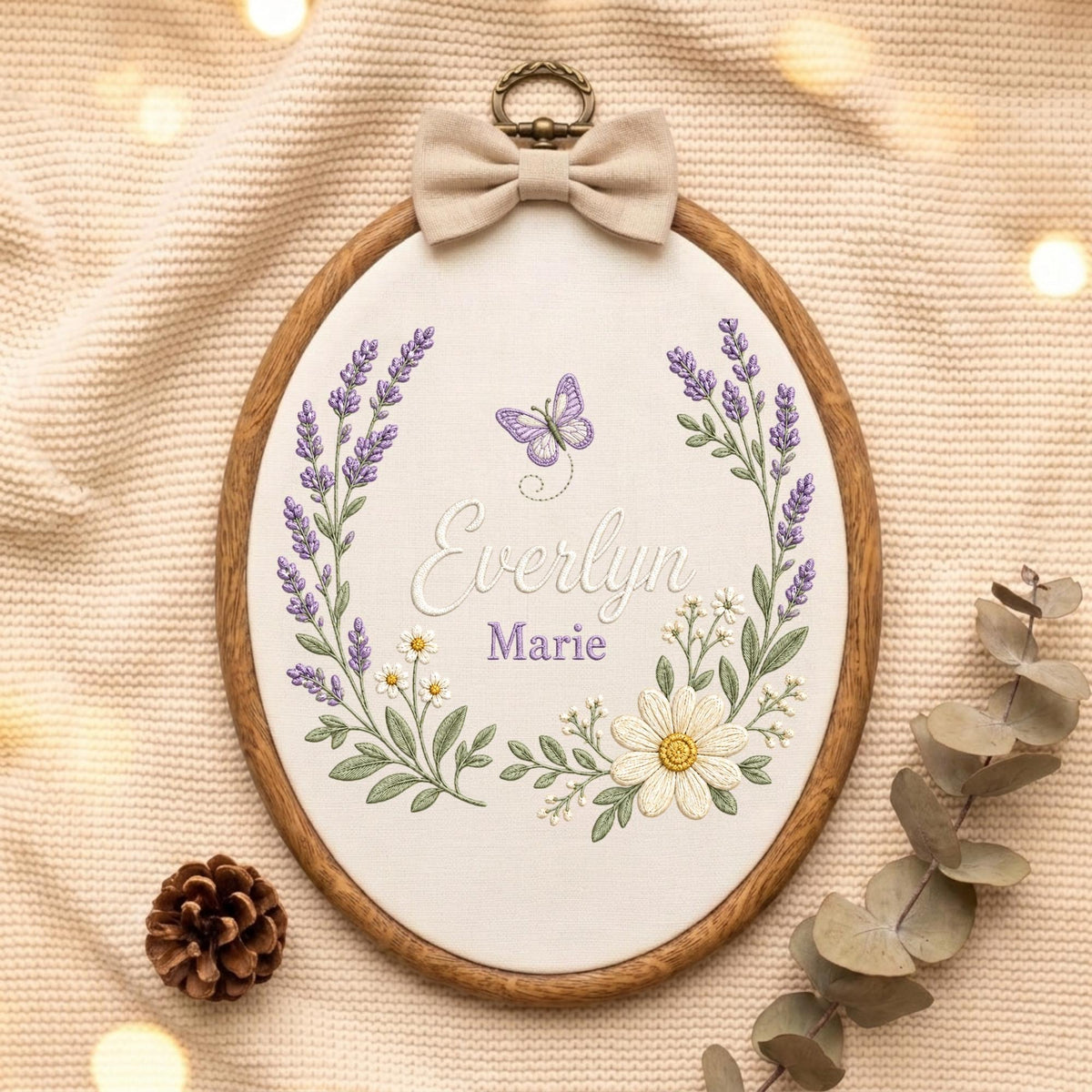 May include: An oval wooden embroidery hoop containing white fabric with a hand-stitched design of lavender sprigs, daisies, and a purple butterfly. The name Everlyn Marie is embroidered in the center, topped with a beige fabric bow and metal ring.