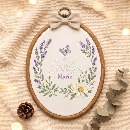 May include: An oval wooden embroidery hoop containing white fabric with a hand-stitched design of lavender sprigs, daisies, and a purple butterfly. The name Everlyn Marie is embroidered in the center, topped with a beige fabric bow and metal ring.