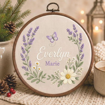 May include: A circular wooden embroidery hoop containing beige fabric with a hand-stitched design. The embroidery features a lavender and daisy wreath surrounding the names Everlyn Marie in white and purple script, with a small purple butterfly.