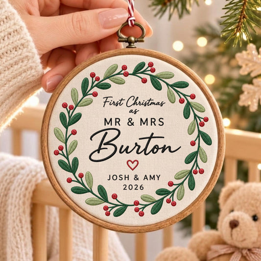May include: A round wooden embroidery hoop ornament featuring a white fabric center with an embroidered green holly wreath and red berries. The text reads First Christmas as MR &amp; MRS Burton, JOSH &amp; AMY, and 2026, with a small red heart icon.