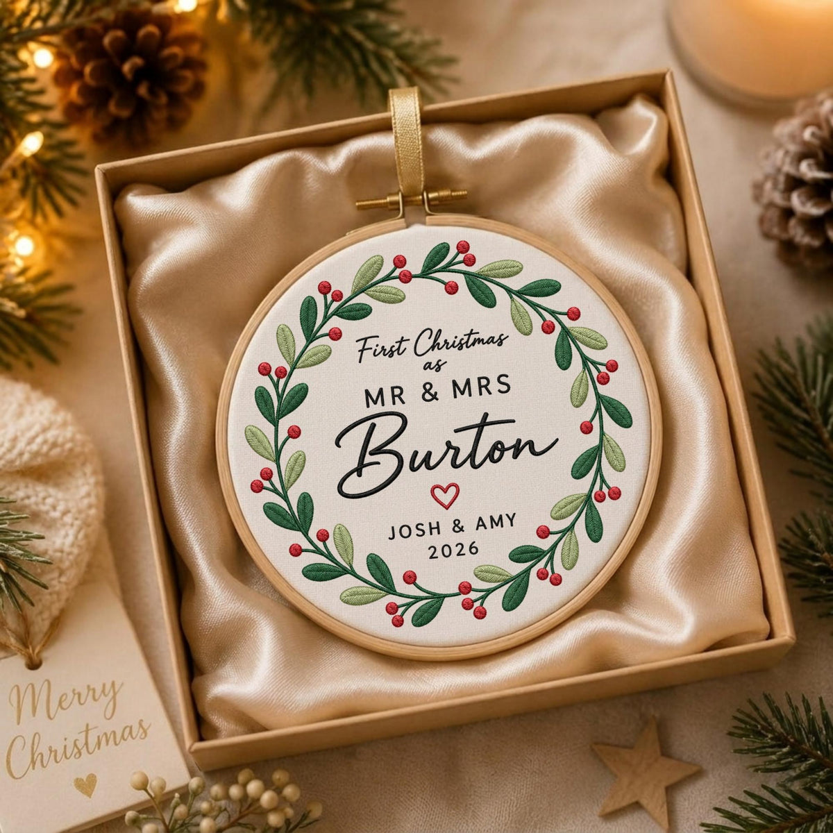 May include: A round embroidery hoop ornament featuring a white fabric center with a green leaf and red berry wreath. The text reads First Christmas as MR &amp; MRS Burton with a small heart, JOSH &amp; AMY, and 2026. It is presented in a gift box.