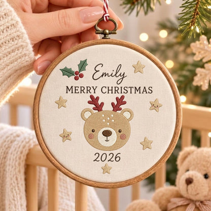 May include: A circular wooden embroidery hoop containing off-white fabric with an embroidered design featuring a brown bear face with red antlers, the name Emily, the text MERRY CHRISTMAS, the year 2026, green holly leaves, and gold stars.