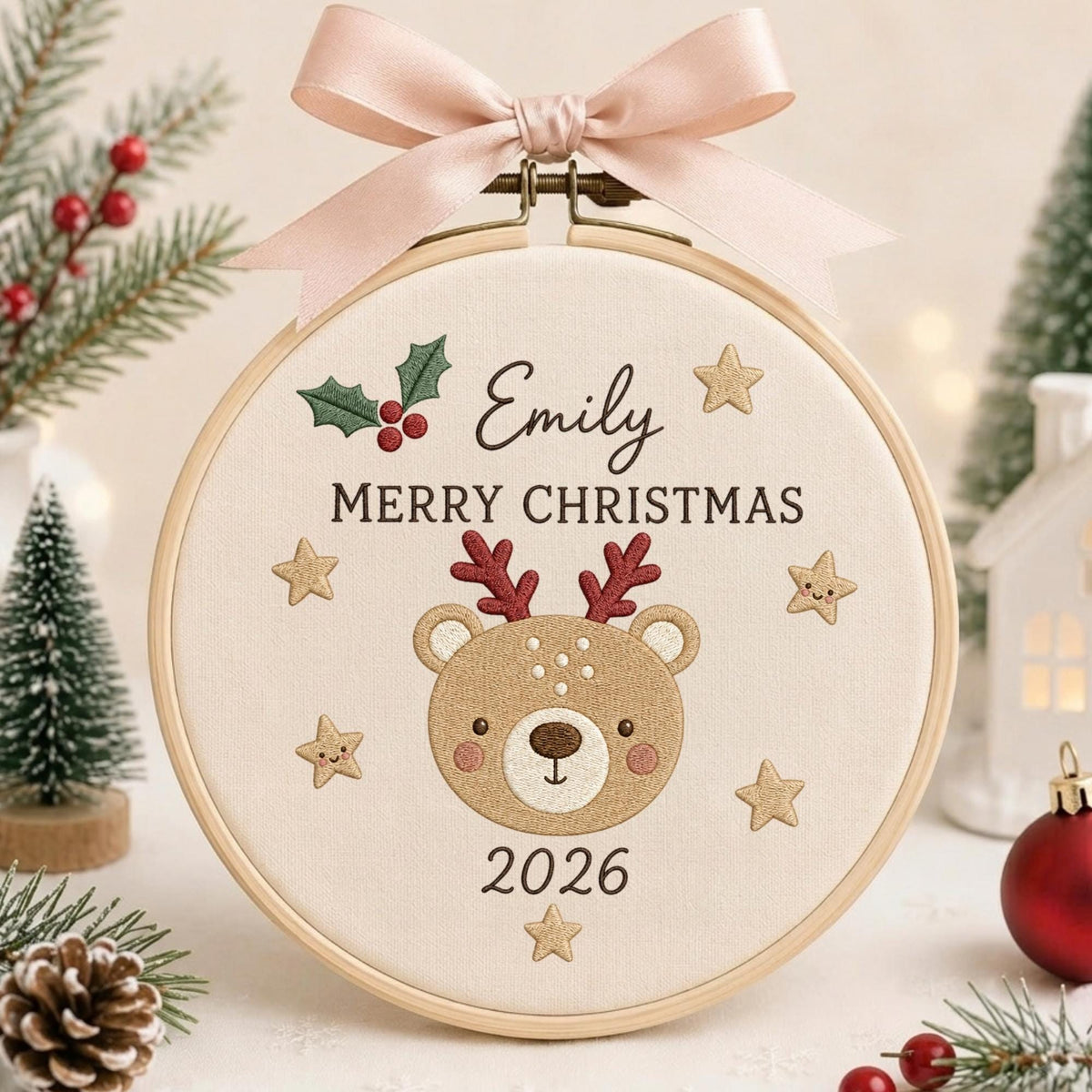 May include: A round wooden embroidery hoop containing cream fabric with an embroidered brown bear face, red antlers, and gold stars. The text reads Emily, MERRY CHRISTMAS, and 2026. A pink ribbon is tied to the top of the hoop above green holly leaves.