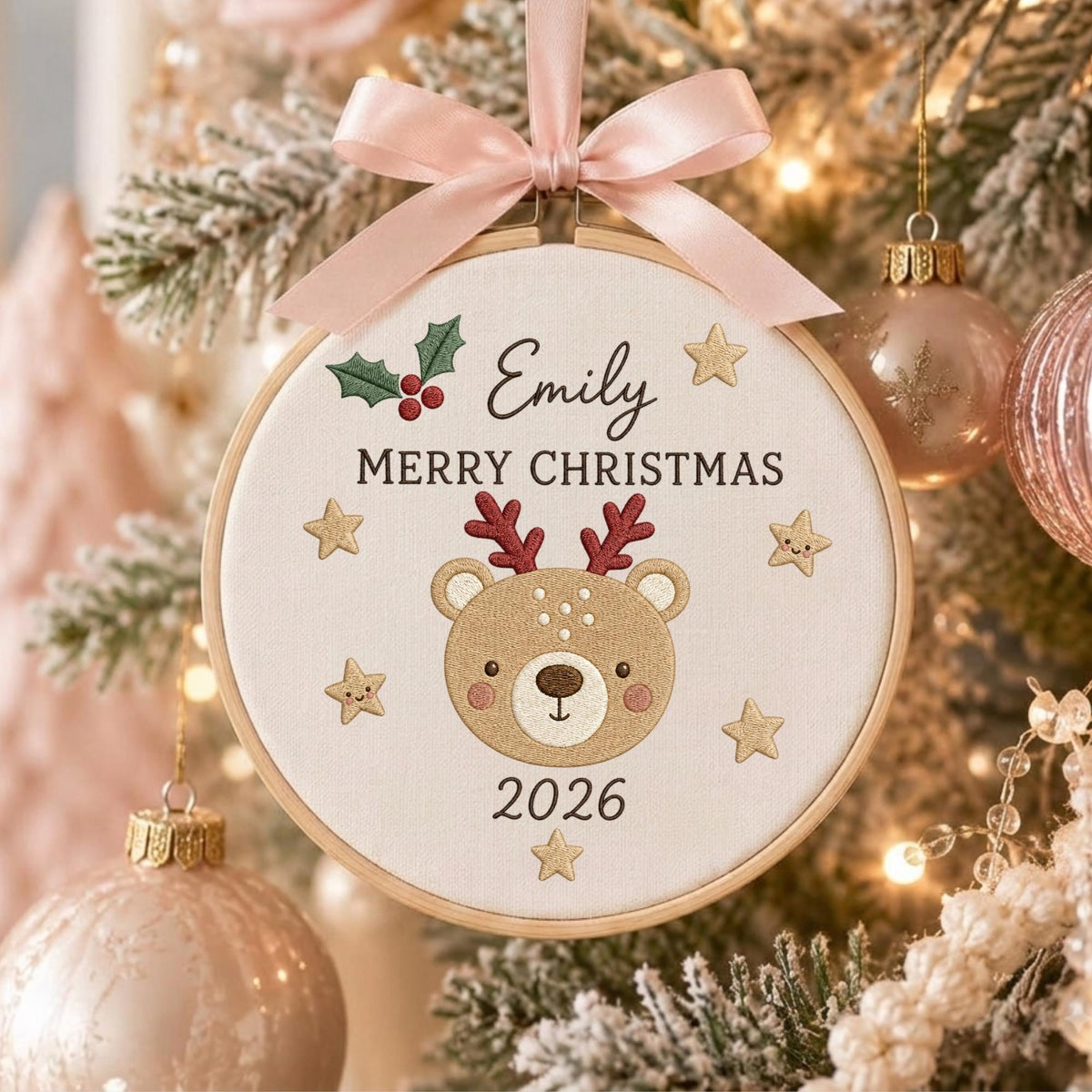 May include: A round wooden embroidery hoop ornament featuring a cream fabric center with a stitched brown bear face wearing red antlers. The text reads Emily, MERRY CHRISTMAS, and 2026, accented by green holly, red berries, and gold stars, topped with a pink ribbon.