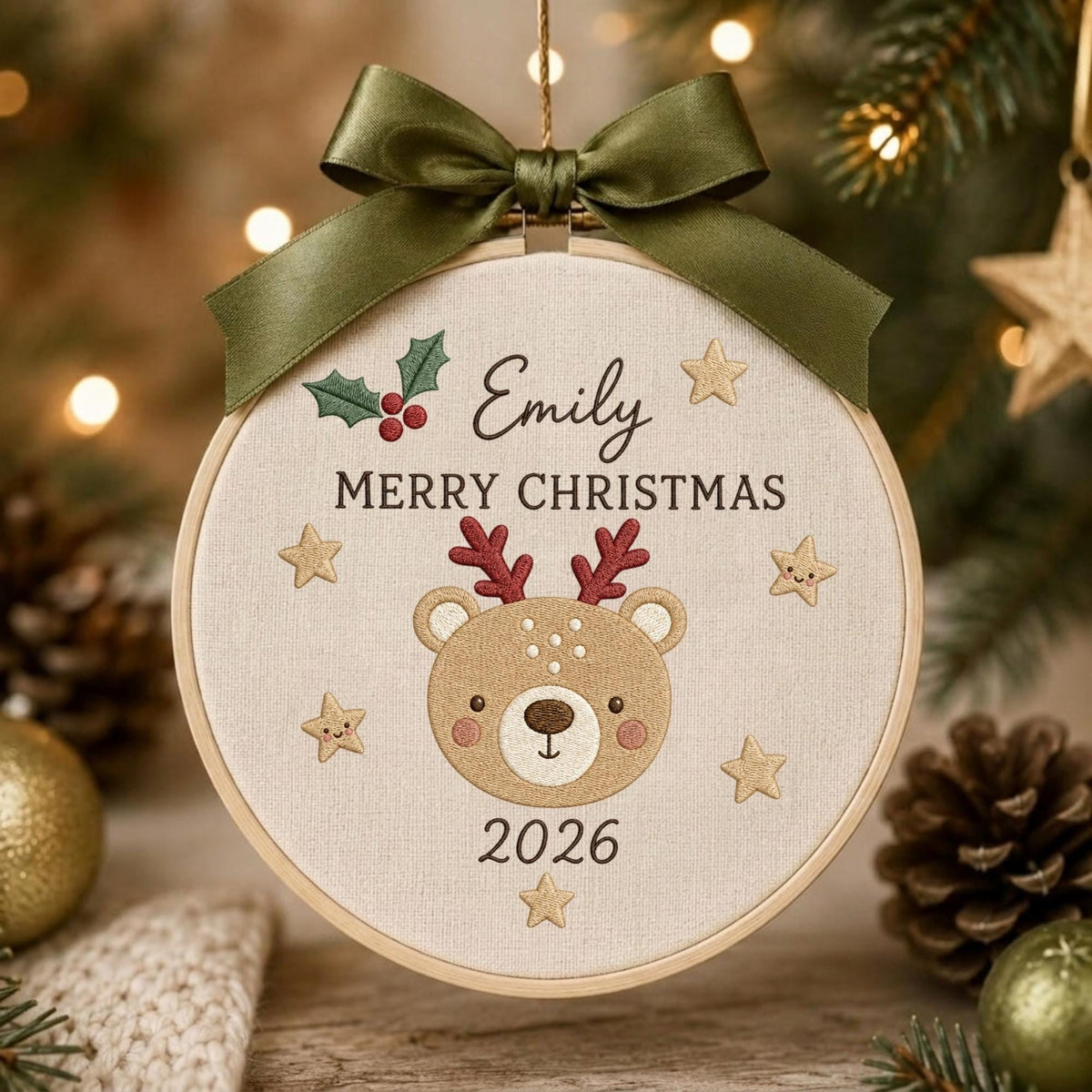 May include: A round embroidery hoop ornament featuring a beige fabric background with the embroidered name Emily, the text MERRY CHRISTMAS, and the year 2026. A central brown bear face with red antlers is surrounded by small gold stars and a green ribbon bow.