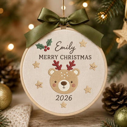 May include: A round embroidery hoop ornament featuring a beige fabric background with the embroidered name Emily, the text MERRY CHRISTMAS, and the year 2026. A central brown bear face with red antlers is surrounded by small gold stars and a green ribbon bow.