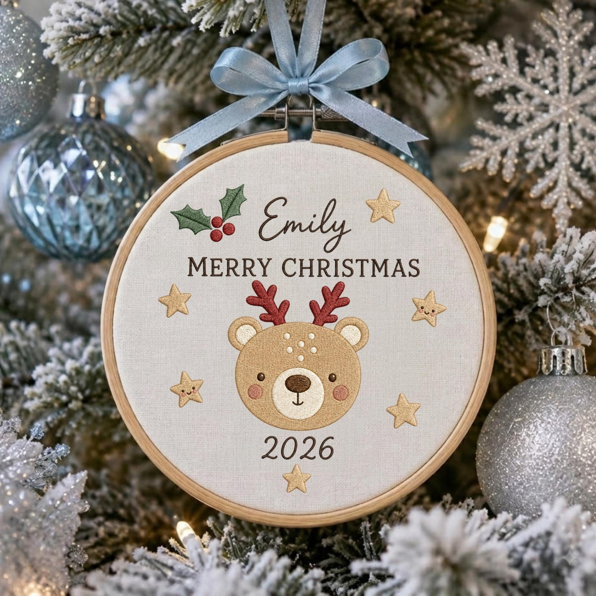 May include: A round wooden embroidery hoop ornament featuring a light beige fabric center with an embroidered brown reindeer face, red antlers, green holly, and gold stars. The text reads Emily, MERRY CHRISTMAS, and 2026. A light blue ribbon bow is attached.