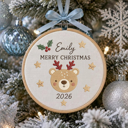 May include: A round wooden embroidery hoop ornament featuring a light beige fabric center with an embroidered brown reindeer face, red antlers, green holly, and gold stars. The text reads Emily, MERRY CHRISTMAS, and 2026. A light blue ribbon bow is attached.