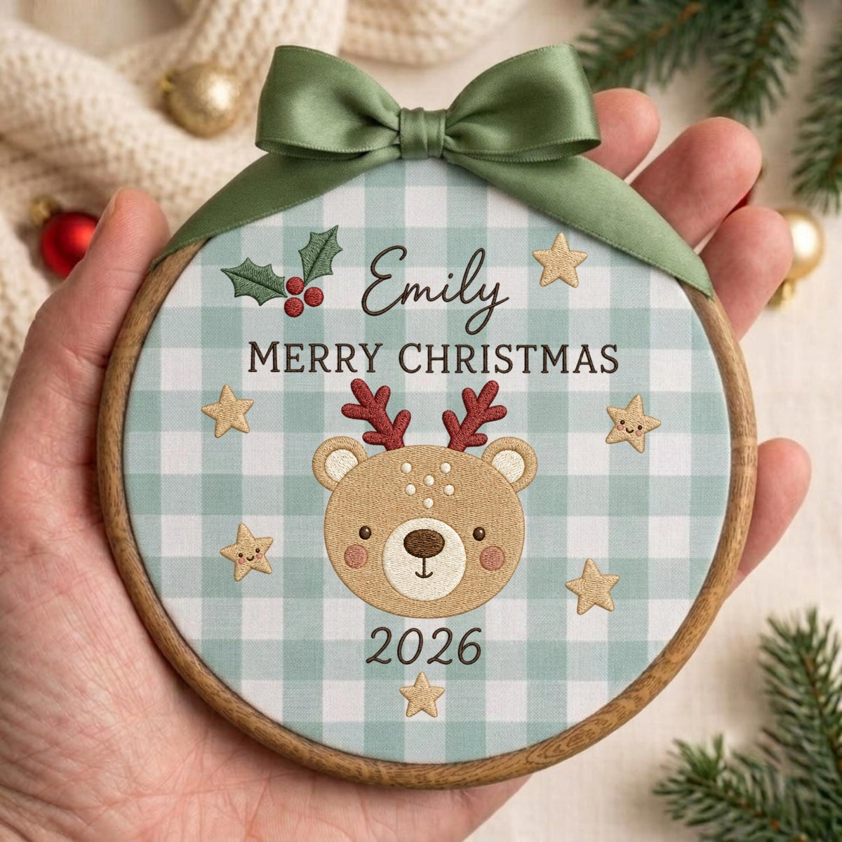 May include: A round wooden embroidery hoop ornament featuring a light blue and white gingham fabric background. It displays the name Emily, the text MERRY CHRISTMAS, the year 2026, a reindeer face, gold stars, and holly, topped with a sage green ribbon.