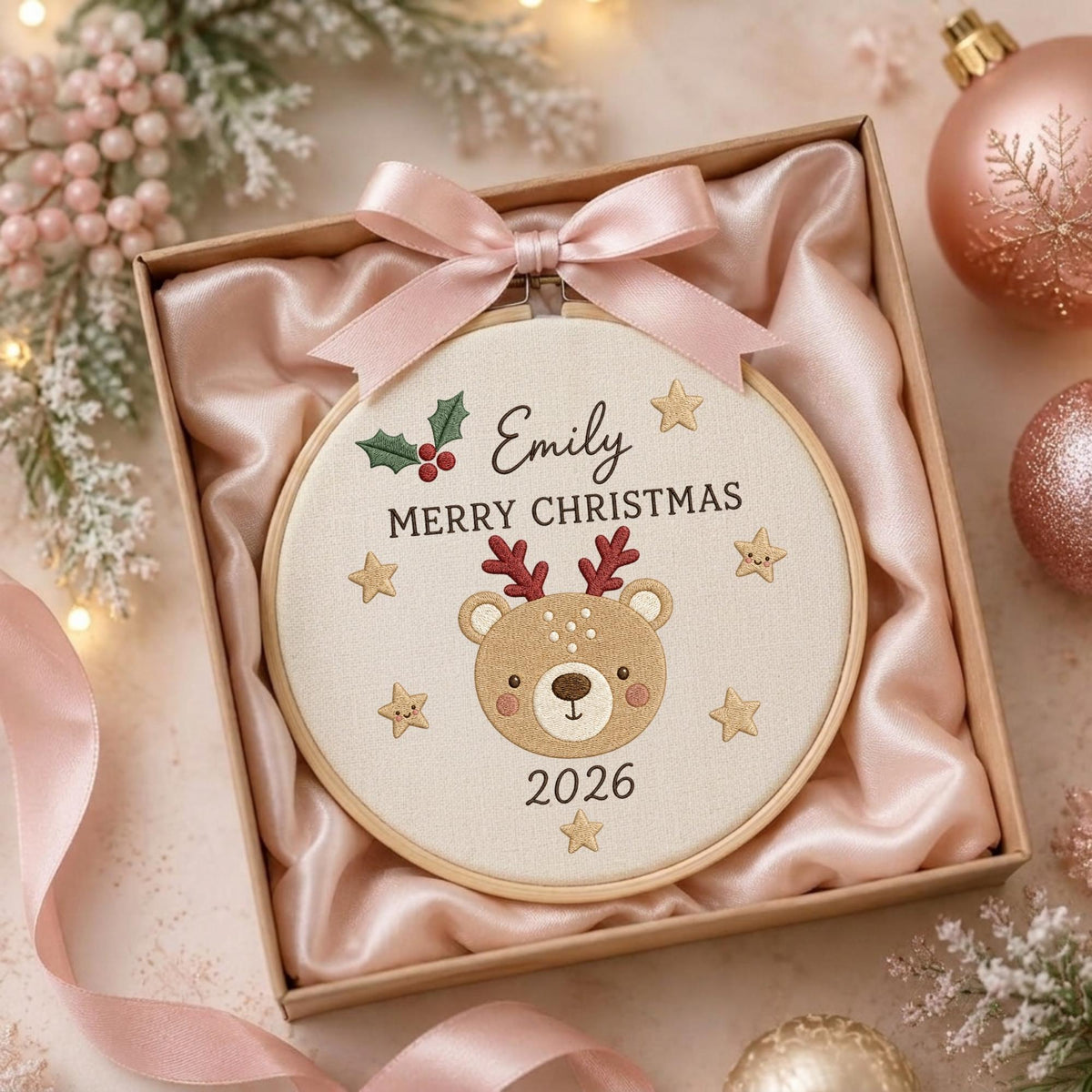 May include: A round wooden embroidery hoop containing a cream-colored fabric with an embroidered brown bear face, red antlers, green holly, gold stars, and the text Emily MERRY CHRISTMAS 2026. A light pink satin ribbon bow is tied at the top.