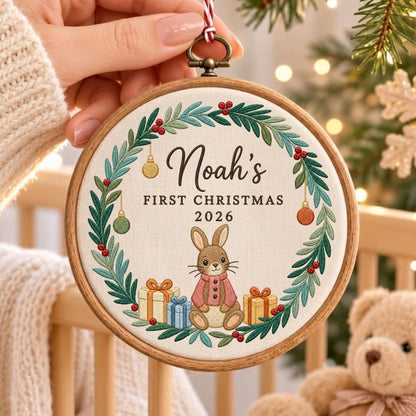 May include: A round wooden embroidery hoop ornament featuring a white fabric center with a green pine wreath, red berries, and hanging ornaments. The text reads Noah's FIRST CHRISTMAS 2026 above a brown rabbit wearing a pink sweater and gift boxes.