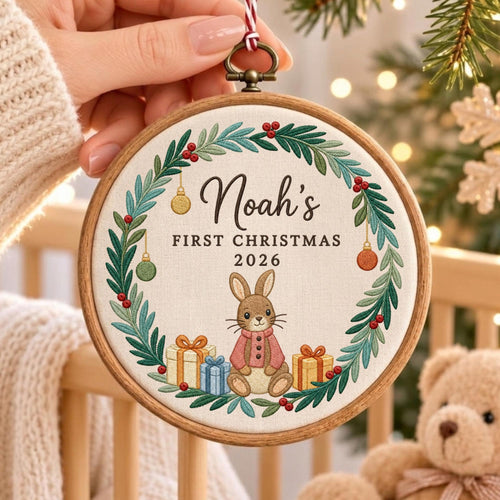 May include: A round wooden embroidery hoop ornament featuring a white fabric center with a green pine wreath, red berries, and hanging ornaments. The text reads Noah's FIRST CHRISTMAS 2026 above a brown rabbit wearing a pink sweater and gift boxes.