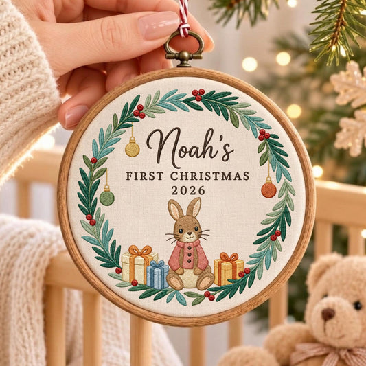 May include: A round wooden embroidery hoop ornament featuring a white fabric center with a green pine wreath, red berries, and hanging ornaments. The text reads Noah's FIRST CHRISTMAS 2026 above a brown rabbit wearing a pink sweater and gift boxes.