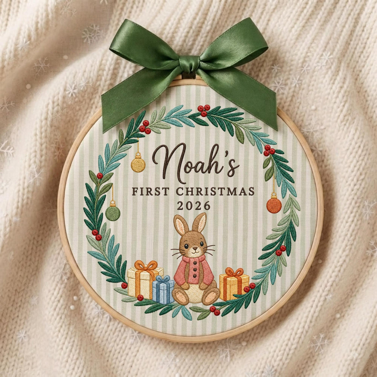 May include: A round wooden embroidery hoop featuring a fabric background with light green vertical stripes. The design includes a wreath of green leaves and red berries, a brown rabbit, gift boxes, and the text Noah's FIRST CHRISTMAS 2026.