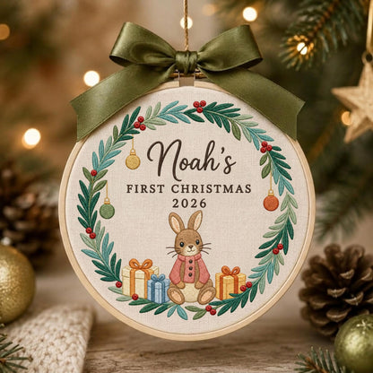 May include: A round embroidery hoop ornament featuring a beige fabric background with a green wreath of leaves and red berries. The text reads Noah's FIRST CHRISTMAS 2026 above a brown rabbit wearing a pink sweater, surrounded by gift boxes.