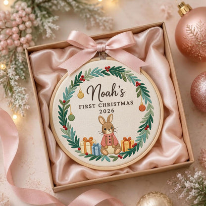 May include: A round wooden embroidery hoop containing fabric with a stitched wreath of green leaves and red berries. The center features a brown rabbit wearing a pink coat, surrounded by gifts, with the text Noah's FIRST CHRISTMAS 2026.