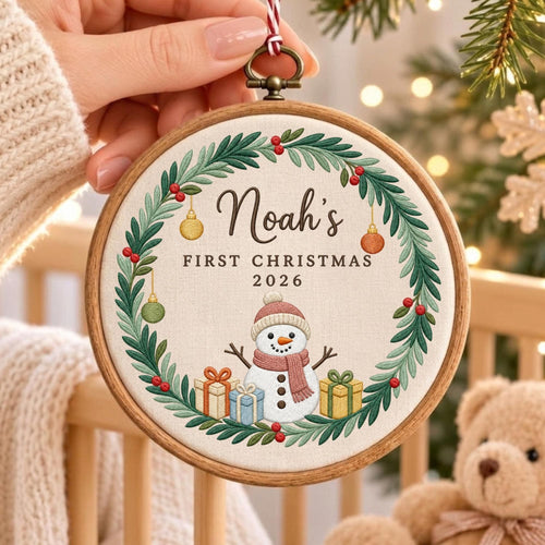 May include: A round wooden embroidery hoop containing a fabric ornament with a green wreath, red berries, and hanging ornaments. The center features a snowman wearing a hat and scarf, surrounded by gift boxes, with text reading Noah's FIRST CHRISTMAS 2026.
