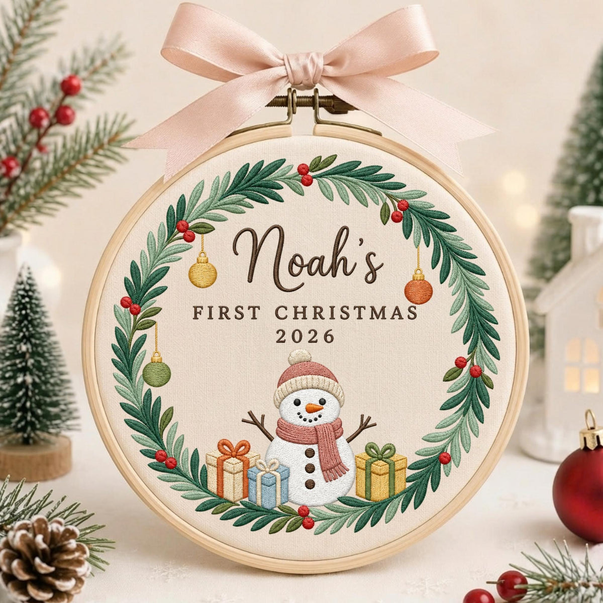 May include: A round wooden embroidery hoop containing fabric with a stitched wreath of green leaves and red berries. The center features a snowman wearing a pink hat and scarf, three gift boxes, and the text Noah's FIRST CHRISTMAS 2026.